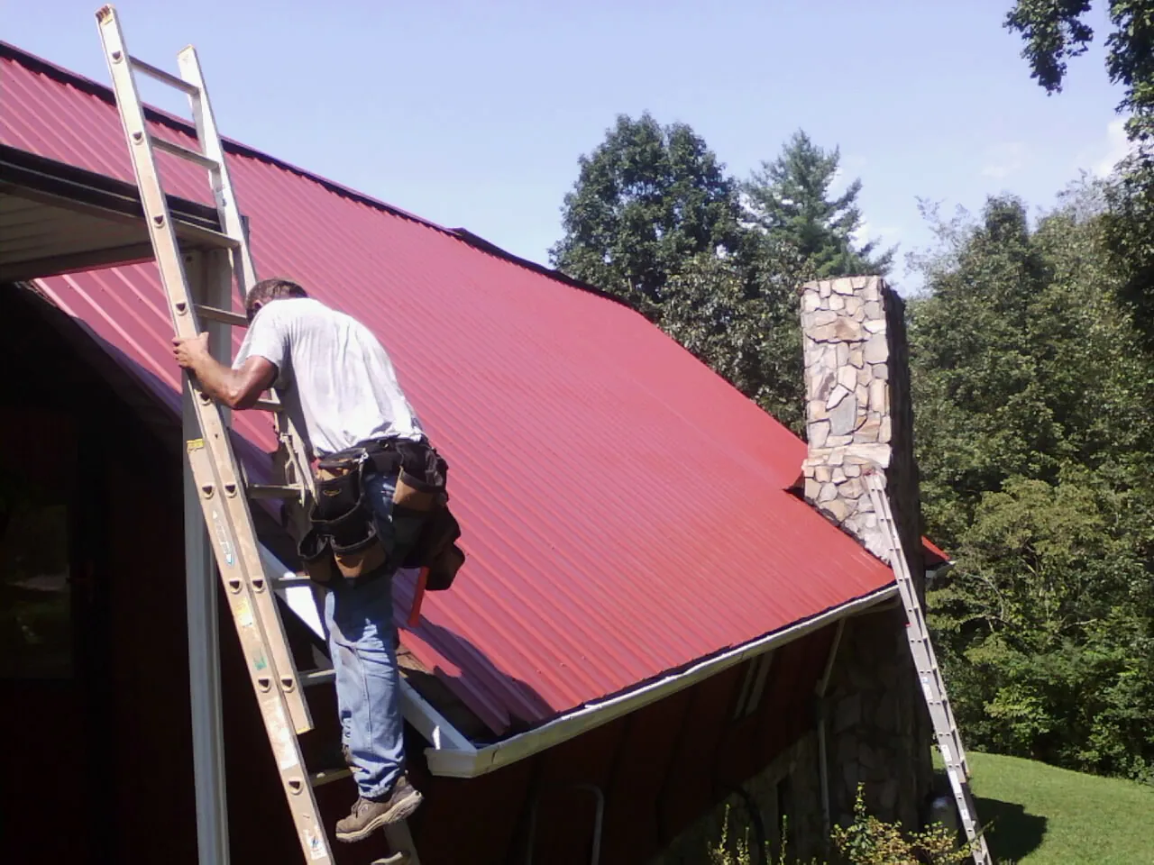 Roofer on a ladder beside a red metal roof and stone chimney — Soffit & Fascia Repair by Roofing Brothers in Cascade Locks